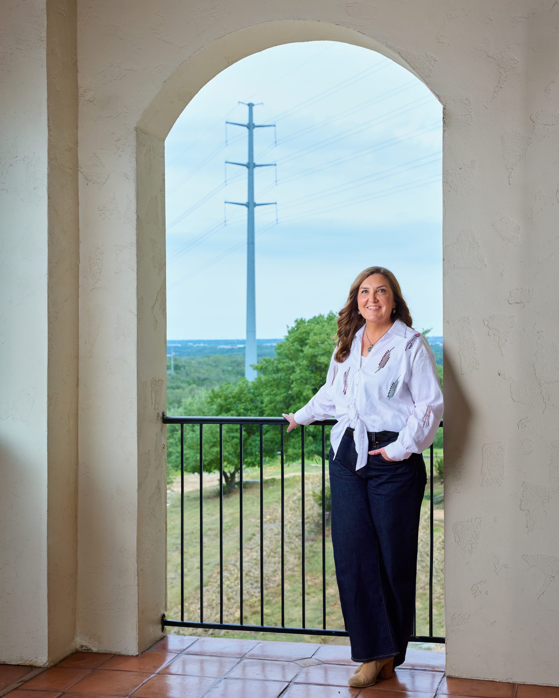 Keri Waters on a balcony framed by a stucco arch, with a high-voltage transmission tower and the Texas Hill Country behind her.